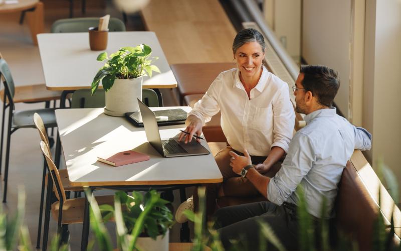 Twee professionals in een cafe samen aan het werk. Je ziet ze van veraf aan tafel zitten. Een vrouw links en een man rechts.