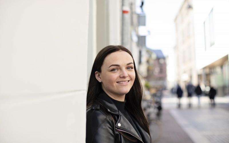 Portret van jonge vrouw. Ze staat op straat tegen een muur geleund, heeft donker haar en een zwart leren jasje aan.