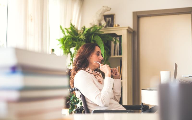 Vrouw in rolstoel zit achter haar bureau. Ze kijkt voor zich uit.