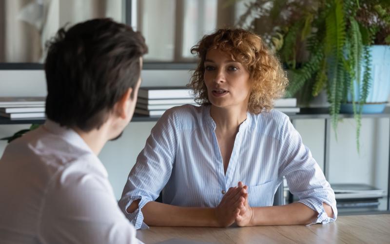 Een man en een vrouw zitten aan tafel en zijn in gesprek. De man is van achteren gefotografeerd en heeft donker haar. De vrouw zie je van voren en heeft rode krullen.