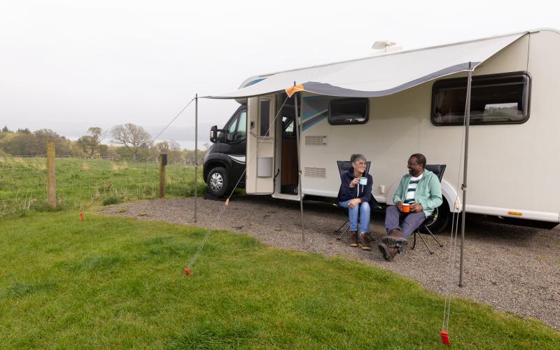 Een vrouw en een man zitten voor een camper op twee campingstoelen. Ze zijn in gesprek.