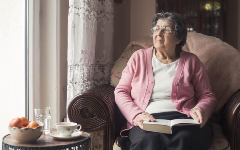 Senior vrouw zit op een stoel bij het raam. Ze heeft een boek vast.