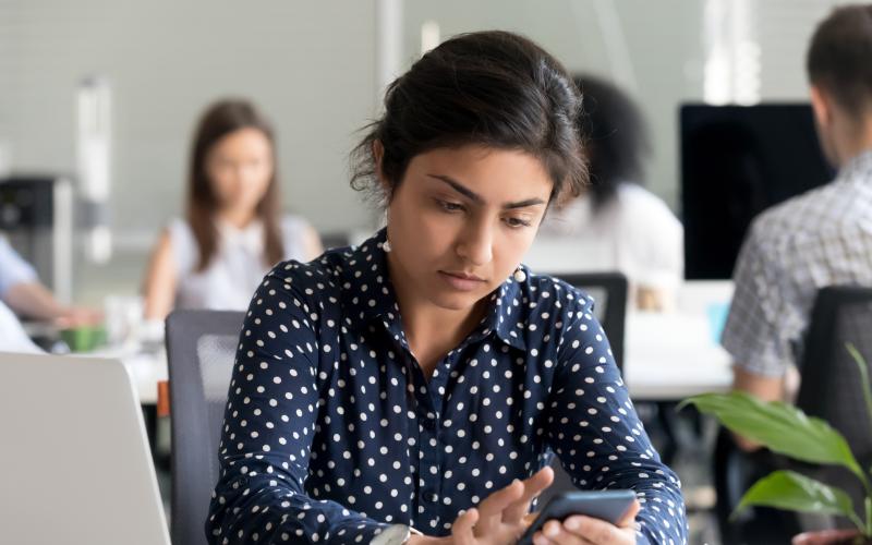 Jongvolwassen vrouw met wit blauw gestipte blouse zit achter laptop met telefoon in de hand