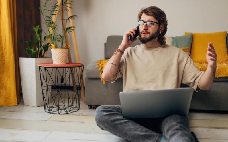 Jongvolwassen man zit op de grond voor zijn bank. Hij is aan het bellen en heeft zijn laptop op schoot.