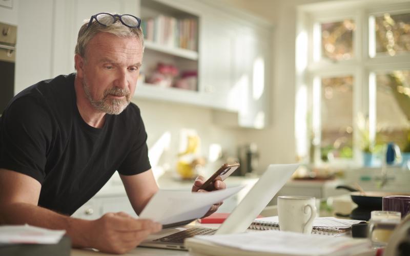 Man staat in zijn keuken en leest een brief. Hij houdt ook zijn telefoon vast.