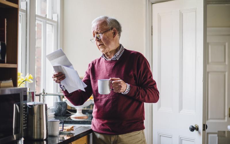 Senior man staat in de keuken met een brief in zijn ene hand en een kopje koffie in zijn andere hand.