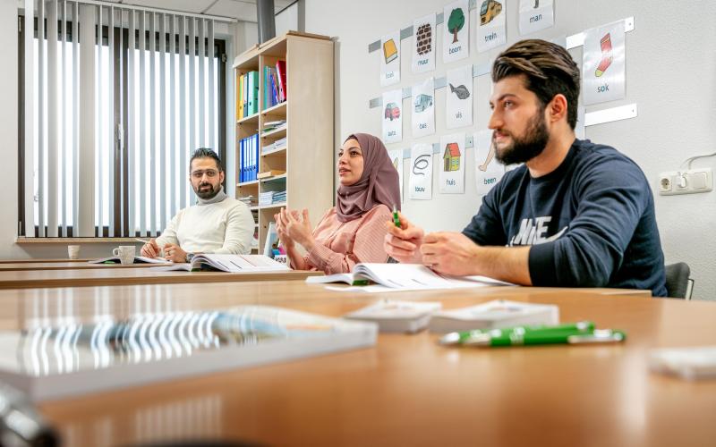 Drie personen zitten naast elkaar in een klaslokaal. Ze volgen een taalles.