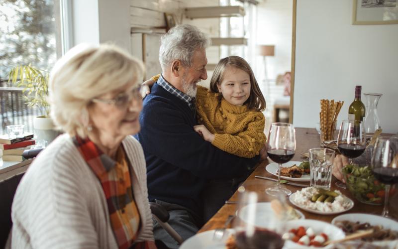 Senior vrouw en man, de grootouders, zitten aan een gedekte tafel met een van de kleinkinderen op schoot.