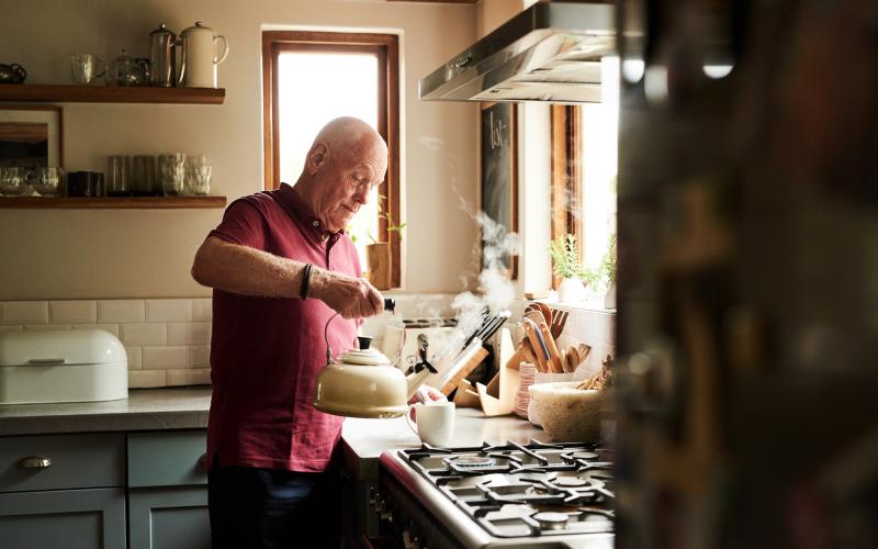 Witte senior man staat in de keuken. Hij heeft een fluitketel vast en schenk heet water in zijn kopje.