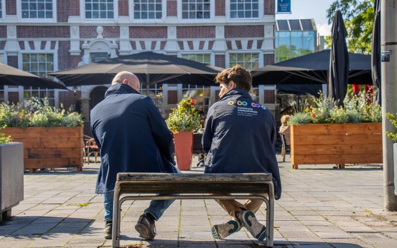 Twee medewerkers van de Nationale ombudsman zitten op een bankje in de zon (gefotografeerd in de rug). 