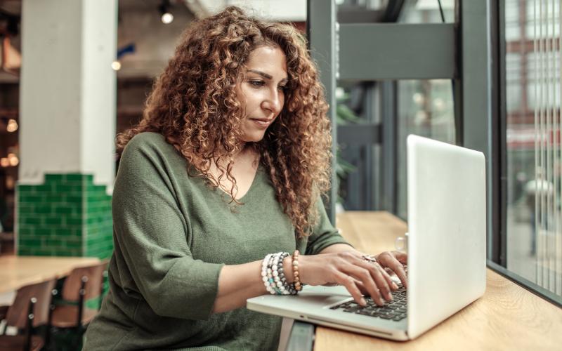 Vrouw met lang krullend haar zit aan een tafel en werkt op haar laptop