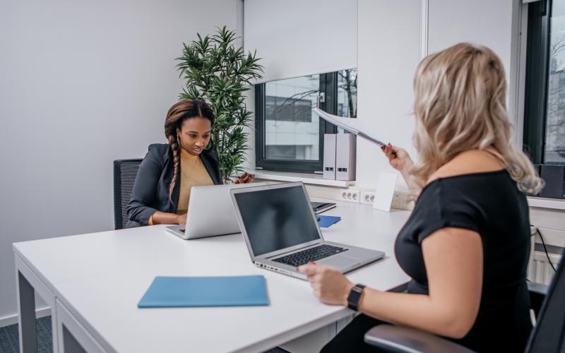 Twee vrouwen aan een bureautafel. Een vrouw overhandigt een stapel papieren aan de andere