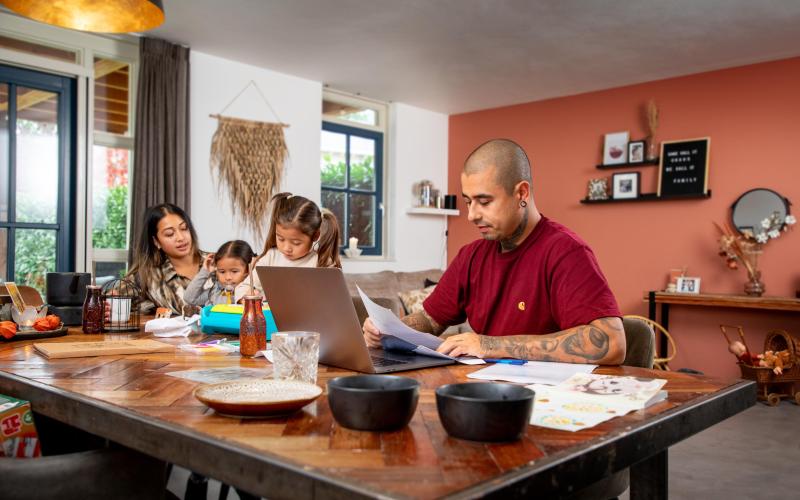 Van links naar rechts: moeder, twee dochtertjes en vader zitten aan tafel. Vader zit achter de laptop en heeft een brief vast.