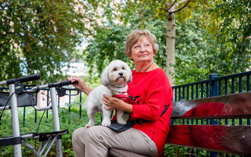 Oudere dame zit op een bankje in het park. Ze heeft haar witte hondje op schoot.