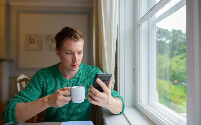 Jongeman zit aan tafel en heeft een kopje koffie en zijn telefoon vast