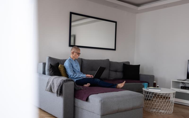 Een vrouw met kaalgeschoren hoofd zit op de bank en heeft een laptop op haar schoot