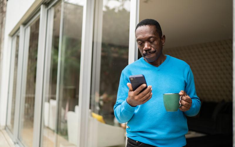 Man staat bij het raam en kijkt op zijn telefoon. In zijn andere hand heeft hij een kopje vast.