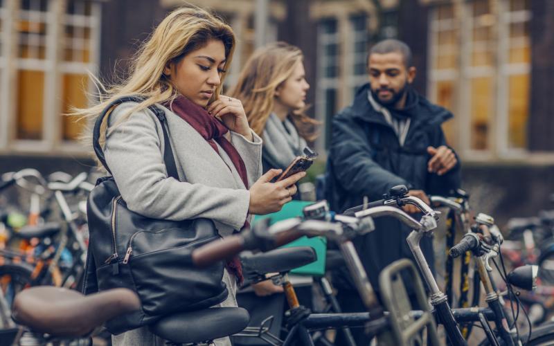 Een jonge student kijkt bezorgd naar haar telefoon. Op de achtergrond staan haar vrienden.