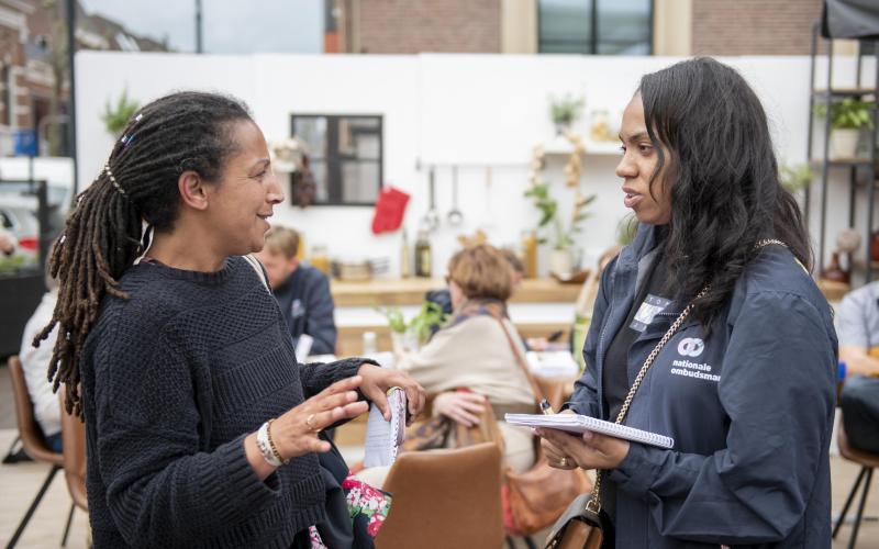 Twee vrouwen staan op een plein voor de pop-upkeuken van de Nationale ombudsman. Ze zijn in gesprek. 