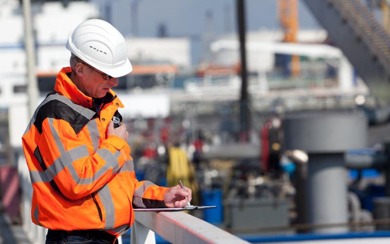 Foto van een man in een oranje veiligheidsjas en met een witte helm op. Hij staat voor een haven.