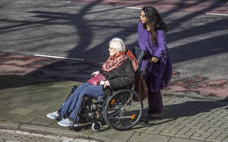 Mantelzorger (vrouw, lang donker haar en paarse jas) duwt rolstoel oudere vrouw (grijs haar, bril, donkere jas en rode sjaal