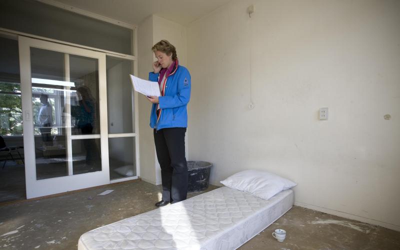 Vrouw met blauwe jas staat in lege kamer met matras op de grond