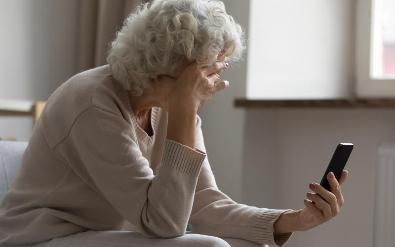 Een oudere vrouw met haar met smartphone in haar hand. Ze leunt met haar hoofd op haar andere hand.