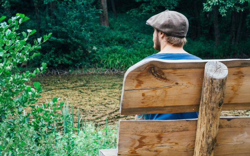 Man zit in de natuur op een bank. Hij is van achter gefotografeerd.