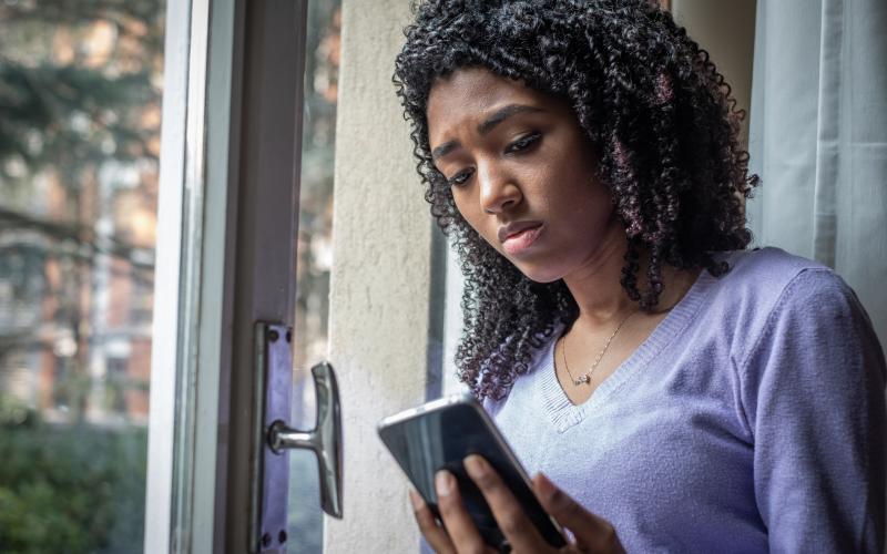 Vrouw kijkt zorgelijk op haar mobiel en staat bij het raam