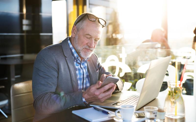 Man op leeftijd zit in café achter zijn laptop met telefoon in zijn hand