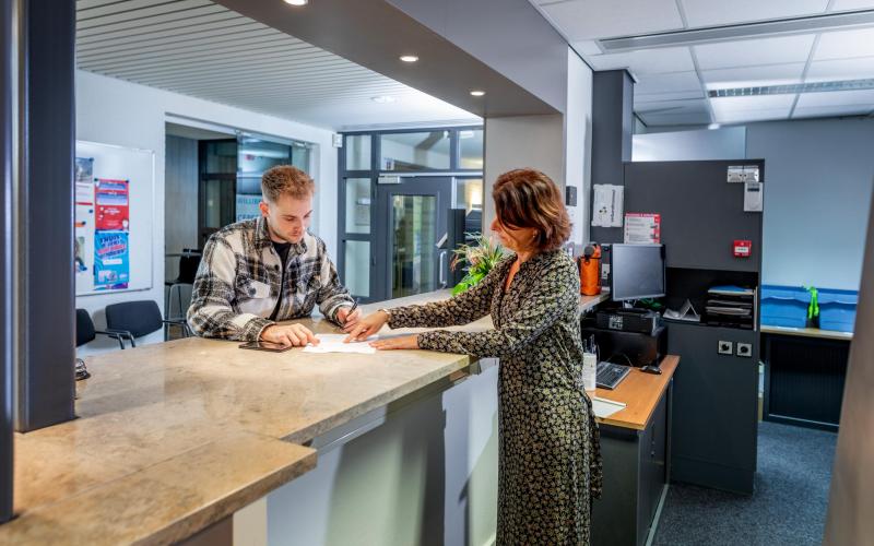 Jongere man aan de balie bij het gemeentehuis. De vrouw achter de balie laat hem een formulier zien waar hij moet tekenen.