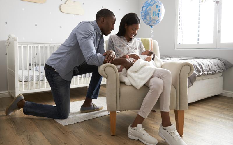 Vrouw zit met baby in haar armen op een stoel. Haar man zit geknield achter haar en kijkt glimlachend naar de baby.