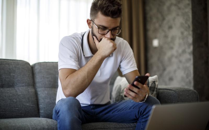 Jongeman met bril en wit t-shirt zit op zijn bank en kijkt ongerust naar zijn telefoon