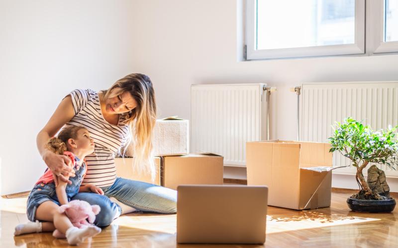 Moeder en kind zitten op de vloer van hun lege appartement van verhuisdozen voor zich.