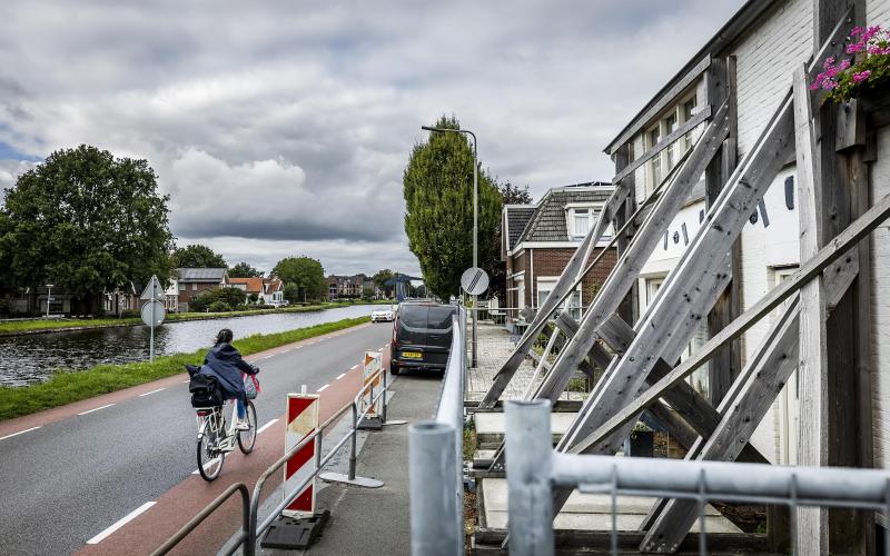 Vrouw fiets langs kanaal. Naast haar staan huizen die ondersteund worden