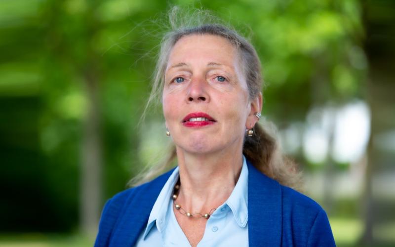 Vrouw (Janny Arends) staat in het park. Haar in een staart, rode lippenstift en draagt blauw jasje 