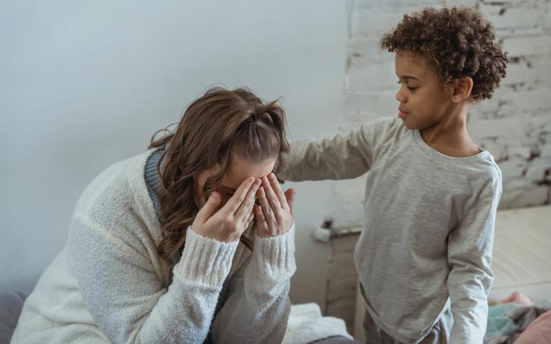 Jonge moeder zit voorover gebogen met handen voor gezicht, kindje troost haar