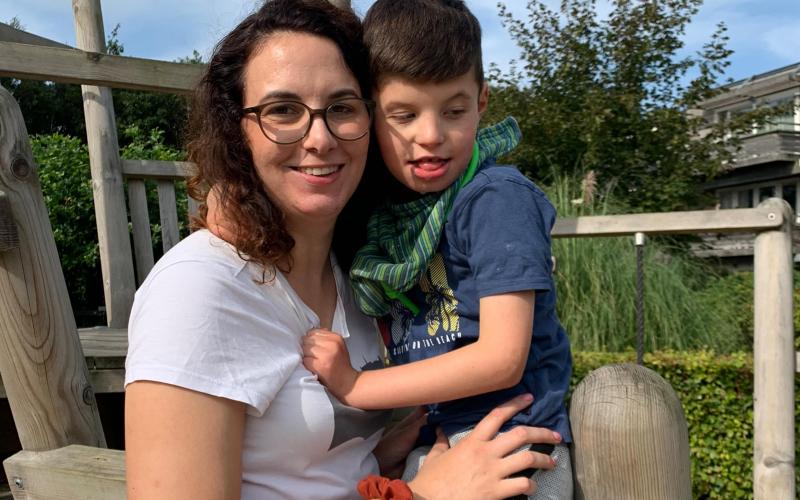 Vrouw met donkerbruin haar en bril kijkt lachend in de camera. Ze houdt haar gehandicapte zoon met donkerbruin haar in haar arm