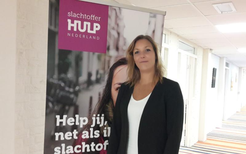 Vrouw (Simone Schuitema) met lang donkerblond haar, draagt wit shirt en zwart jasje. Ze staat in een gang met een gestreepte vloer