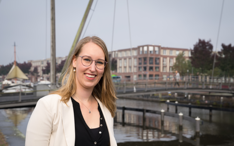 Vrouw (Michelle Koelewijn) heeft blond haar en draagt een bril. Ze staat buiten bij het water (haven)