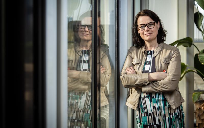 Vrouw met donker halflang haar. Ze draagt een donkere bril, gestreept shirt en beige jasje. Achter haar staat een plant