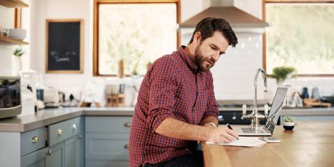 Man zit aan tafel in de keuken en is zaken aan het regelen op zijn laptop. Hij schrijft op een papiertje.
