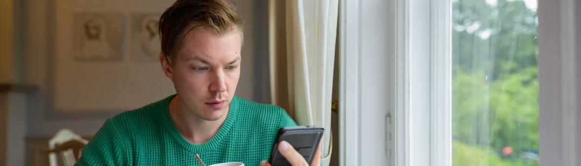 Jongeman zit aan tafel en heeft een kopje koffie en zijn telefoon vast