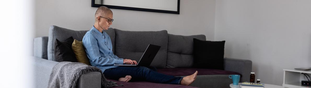 Een vrouw met kaalgeschoren hoofd zit op de bank en heeft een laptop op haar schoot